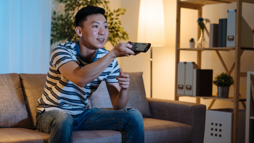 Man sitting on a couch watching TV with a remote control in his hand, pointed at the TV