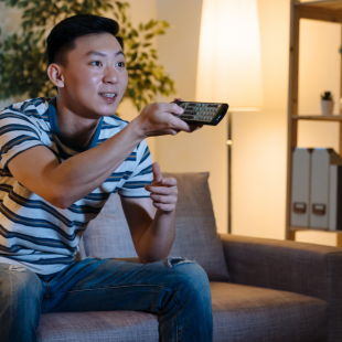 Man sitting on a couch watching TV with a remote control in his hand, pointed at the TV