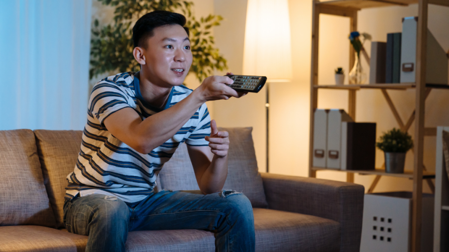 Man sitting on a couch watching TV with a remote control in his hand, pointed at the TV